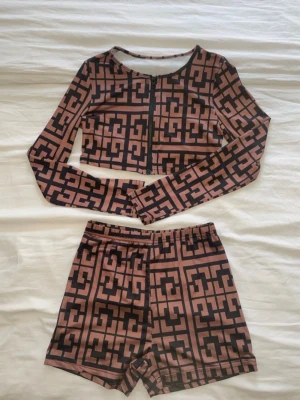 Brun och svart croppad topp och shorts SET från SHEIN - Säljer en croppad långärmad topp från SHEIN i brun och svart med grafiskt rutigt mönster. Toppen har dragkedja framtill och är tillverkad i stretchigt syntetmaterial. Perfekt att matcha med höga byxor eller kjol för en cool look.