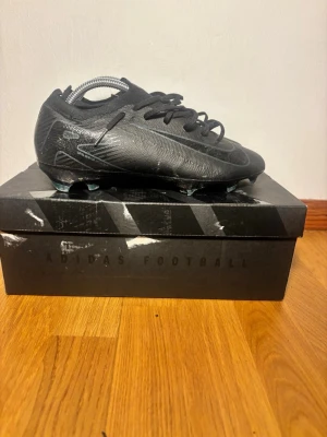 Svarta Nike fotbollsskor med snörning - Säljer ett par svarta Nike fotbollsskor med snygga grafiska detaljer och snörning. Skorna har låg profil och platt sula med dobbar för gräsplan. Ovandelen är tillverkad i syntetmaterial med textilinslag för extra komfort och passform.
