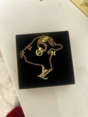 Guldig  halsband - Säljer ett snyggt halsband i guldton med -logga täckt av små glittrande stenar. Kedjan har även en liten blomdetalj -märke vid låset. Perfekt accessoar för att lyfta din outfit.