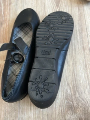 Svarta Mary Jane ballerinaskor med plattform - Säljer ett par svarta Mary Jane-inspirerade ballerinaskor från Flexi med bred plattformssula och rund tå. Skorna har ett justerbart spänne och insida med rutigt foder. Tillverkade i syntetmaterial med robust sula för extra komfort.