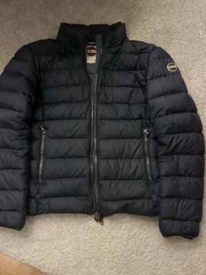 Svart pufferjacka från Colmar - Säljer en svart pufferjacka från Colmar med klassisk quiltad design och två dragkedjefickor framtill. Jackan har hög krage, Colmar-logga på ärmen och är tillverkad i syntetmaterial. Perfekt för kyliga dagar och riktigt snygg till streetwear.