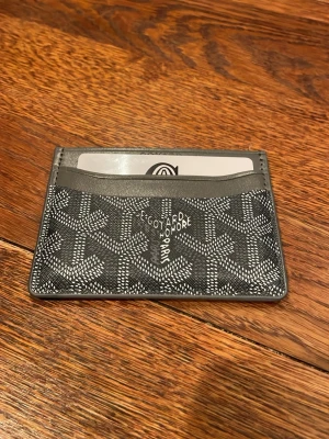 Grå Goy4rd plånbok med mönster - Säljer en stilren grå plånbok från Goy4rd med deras ikoniska grafiska mönster i vitt. Plånboken är tunn och smidig, perfekt för kort och sedlar. Kommer i original grön ask med dustbag. Lyxig accessoar för dig som gillar exklusiva detaljer.