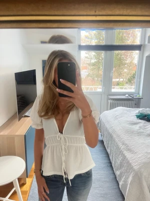 Vit blus med snörning och puffärm - Säljer en vit blus i lätt strukturerad bomull med korta puffärmar och djup v-ringning. Blusen har snörning framtill i midjan och en lätt volang nedtill.