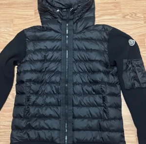 Svart pufferjacka från Moncler - Svart pufferjacka från Moncler med huva och ribbade stickade ärmar. Jackan har quiltad front, dragkedja och en ficka med dragkedja på ärmen. Moncler-logga på vänster ärm. Perfekt för dig som gillar stilren och sportig look.