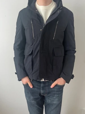 Svart vindjacka från Herno med huva - Svart vindjacka från Herno med huva, dragkedja och tryckknappar framtill. Jackan har två stora fickor och justerbara detaljer vid ärmslut. Materialet är lätt och känns som polyester, perfekt för blåsiga dagar. Snygg och stilren design med Herno-logga på knapparna.