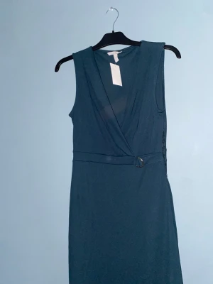 Mörkblå omlottklänning från H&M - Snygg mörkblå omlottklänning från H&M i storlek S. Klänningen är ärmlös med v-ringad omlottdesign och ett bälte i midjan med metallspänne. Perfekt för dig som gillar enkel men stilren look.