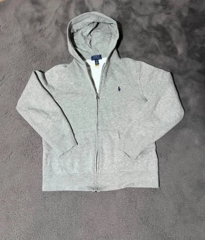Grå hoodie från Polo Ralph Lauren - Snygg grå hoodie från Polo Ralph Lauren med dragkedja och huva. Tröjan har långa ärmar, ribbade muddar och en klassisk Polo-logga broderad på bröstet. Perfekt för vintern och enkel att matcha med jeans❄️ Storlek L(14-16). Sitter som XS