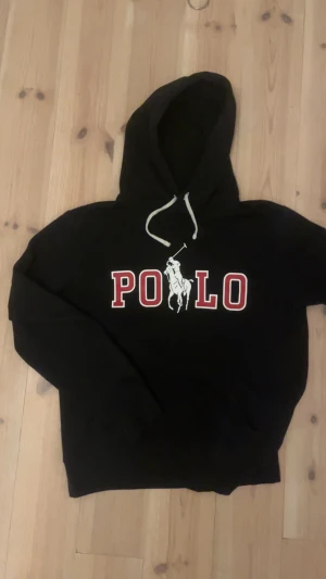 Svart hoodie från Polo Ralph Lauren - Svart hoodie från Polo Ralph Lauren med stor vit och röd POLO-logga och polospelare på bröstet. Tröjan har huva med vita snören och lång ärm. Perfekt för dig som gillar klassisk streetstyle och vill ha en snygg logotröja.