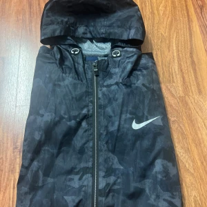 Nike svart camo vindjacka med huva - Nike vindjacka i svart med diskret camouflagemönster och reflekterande logga på bröstet. Jackan har huva med dragsko, hel dragkedja framtill och två sidofickor med dragkedja. Lätt och smidig, perfekt för blåsiga dagar.