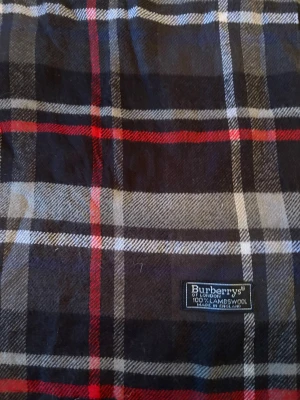 Rutig ullhalsduk från Burberrys - Snygg halsduk från Burberrys i klassiskt rutig design med svarta, gråa, vita och röda toner. Gjord av 100% lammull och har fransar i kanterna. Perfekt för att ge din outfit en cool och tidlös touch.