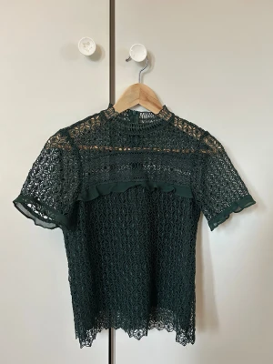 Grön spetsblus från Zara Woman - Säljer en mörkgrön blus från Zara Woman i storlek XS. Blusen är helspets med korta ärmar, hög hals och fina volangdetaljer framtill. Den har ett unikt geometriskt spetsmönster och stängs med dragkedja i nacken. 