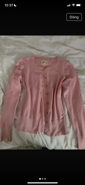 Ljusrosa kofta från Holly & White XS - Supersöt ljusrosa kofta från Holly & White i storlek XS. Koftan har rund hals, långa ärmar och knappar framtill i samma färg som plagget. Mjuk och tunn stickad kvalitet som känns skön mot huden. Perfekt att slänga över en topp för extra stilpoäng.