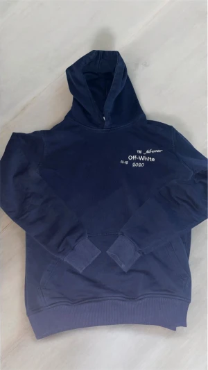Marinblå Off-White hoodie med broderi - Säljer en marinblå hoodie från Off-White med broderad logga och text på bröstet samt stort broderat Off-White-motiv i rött, gult, blått och vitt på ryggen. Tröjan har huva, ribbade muddar och klassisk känguruficka. Perfekt för dig som gillar streetwear.