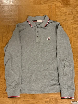Grå långärmad pikétröja Moncler - Snygg grå långärmad pikétröja från Moncler med klassisk krage och knäppning framtill. Nypris 3900kr, köpt på NK i Stockholm. Använd ca 5 gånger. Tröjan har Moncler-logga på bröstet och detaljer i rött och vitt på krage och ärmslut. 