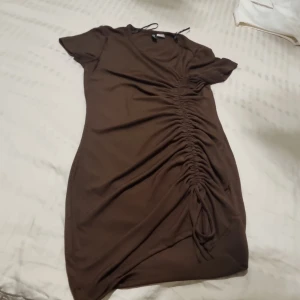 Brun kortklänning med snörning från H&M - Snygg brun kortklänning från H&M Divided i storlek S. Klänningen har korta ärmar och cool snörningsdetalj längs sidan som ger en rynkad effekt. Mjuk och stretchig i materialet, perfekt för dig som gillar en slim passform och enkel stil.