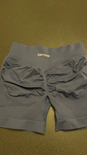  Blå, gråa träningstights shorts från DFYNE - Säljer ett par blå, gråa seamless träningstights shorts från DFYNE. De är högmidjade med bred ribbad linning och har scrunch-detalj bak för extra form. Materialet är stretchigt och passar perfekt för gym eller annan träning. Logga i vitt framtill och baktill på linningen.
