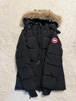 Svart dunjacka från Canada Goose - Säljer en svart dunjacka från Canada Goose med ikonisk patch på ärmen och stor huva med fluffig päls. Jackan har dragkedja framtill, ribbade muddar och flera praktiska fickor. Perfekt för kalla vinterdagar och riktigt snygg streetstil.