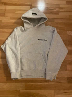 Grå Essentials Fear of God hoodie - Säljer en ljusgrå hoodie från Essentials Fear of God i storlek S. Tröjan har svart logga tryckt både fram och bak, stor huva och klassisk känguruficka. Mjuk insida och ribbade muddar för extra komfort. Perfekt för en chill och stilren look.