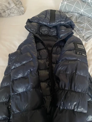 Mörkblå Peak Performance dunjacka - Säljer en mörkblå dunjacka från Peak Performance med glansig finish och huva. Jackan har en snygg quiltad design, dragkedja framtill och logotyp på ärmen. Perfekt för kalla dagar och har en modern, sportig look.