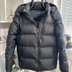 Svart pufferjacka från Polo Ralph Lauren - Svart pufferjacka från Polo Ralph Lauren med röd broderad logga på bröstet. Jackan har huva, flera dragkedjor och quiltad design. Materialet är polyester och jackan har en normal passform med långa ärmar. Perfekt för kalla dagar.