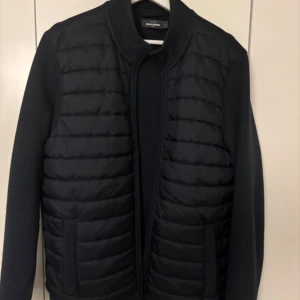 Svart quiltad pufferjacka - Snygg svart pufferjacka med quiltad framsida och släta ärmar och rygg. Jackan har ståkrage, två fickor fram och dragkedja. Perfekt för dig som gillar stilren och modern look.