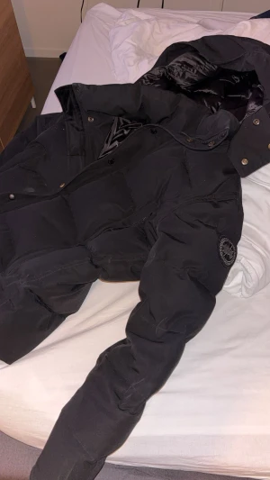 Canada goose  - Säljer en svart pufferjacka från Canada Goose med stor huva och logotyp på ärmen. Jackan har både dragkedja och knappar framtill, samt flera fickor. Perfekt för kalla dagar och har en riktigt schysst, oversized look.