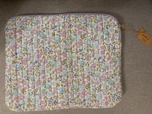 Superfin Blommig quiltad datorväska från Blomz - Superfin quiltad datorväska från Blomz med blommigt mönster i pastellfärger. Väskan har dragkedja och är i mjuk bomull med lila, blå och gröna detaljer. Perfekt för att skydda din laptop med stil.