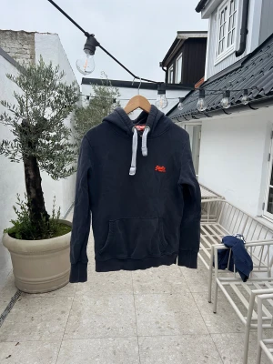 Mörkblå hoodie från Superdry - Snygg mörkblå hoodie från Superdry med grå snörning och röd broderad logga på bröstet. Tröjan har huva, känguruficka och ribbade muddar vid ärmslut och nederkant. Perfekt för chill dagar och enkel att matcha med jeans eller joggers.
