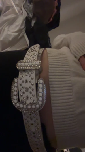 Vitt skärp med silverdetaljer och nitar - Säljer ett vitt bälte med stora silverfärgade spännen och glittriga nitar över hela remmen. Bältet har en bred form och cool western-inspirerad look med dekorativa detaljer runt spännet. Perfekt för att ge outfiten extra edge.