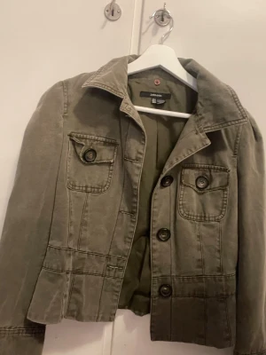 Olivgrön jeansjacka från Zara Basic - Snygg olivgrön jeansjacka från Zara Basic i 100% bomull. Jackan har klassisk krage, två bröstfickor med knappar och markerade sömmar för en cool look. Perfekt att slänga över en hoodie eller t-shirt. Stängs med stora knappar framtill.