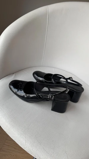 Svarta lackade Mary Jane pumps - Snygga svarta Mary Jane pumps med glansig lackad finish och rund tå. Skorna har en bred låg klack och smala remmar med silverfärgat spänne över foten. Perfekta för dig som gillar en klassisk men trendig look.
