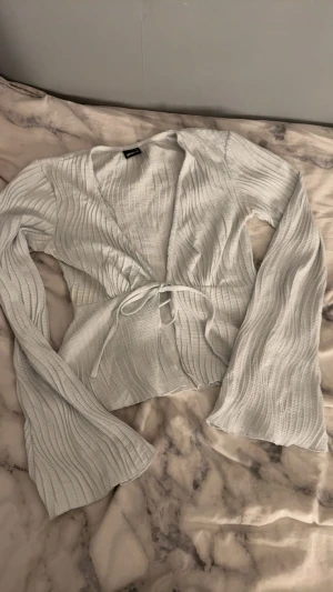 Ljusgrå ribbad omlottopp från Gina Tricot - Säljer en ljusgrå ribbad omlottopp från Gina Tricot med knyt framtill. Toppen har långärmade, lätt utsvängda ärmar och en croppad passform. Materialet är mjukt och stretchigt, perfekt för att styla med jeans eller kjol.