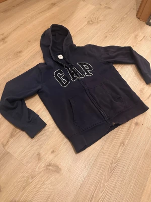 Mörkblå hoodie från GAP med dragkedja - Säljer en mörkblå hoodie från GAP med stor vit logga på bröstet. Tröjan har huva med snörning, två fickor fram och hel dragkedja. Perfekt för chill dagar och enkel att matcha med jeans eller joggers.