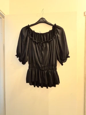 Svart blus med volang och puffärm - Säljer en svart blus med volangkant vid halsen och nederdelen, puffiga korta ärmar och smockad midja. Blusen har en söt knytning framtill och är tillverkad i ett mjukt, lätt material som ger ett flowy intryck. Perfekt för dig som gillar romantiska detaljer.