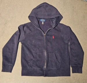 Mörkblå hoodie från Polo Ralph Lauren - Mörkblå hoodie från Polo Ralph Lauren med dragkedja framtill och klassisk huva. Tröjan har en röd broderad logga på bröstet och två fickor framtill. Perfekt för en chill och stilren look.