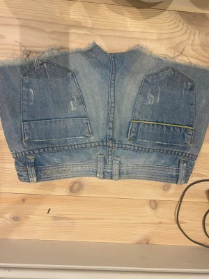 Blå denim tubtopp från Kenzo Jeans - Säljer en unik tubtopp gjord av återvunnen blå denim från Kenzo Jeans. Toppen har råa, fransiga kanter och coola detaljer som bälteshällor, fickor och gul söm. Perfekt för dig som gillar statement-plagg och streetstyle. Passar dig som vill sticka ut!
