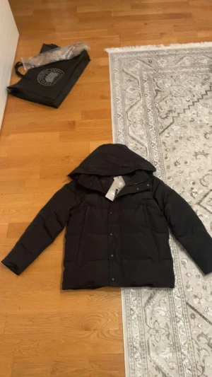  Canada Goose black label - Svart pufferjacka från Canada Goose i storlek XL Fusion. Jackan har huva, långa ärmar och snyggt ton-i-ton broderat märke på ärmen. Materialet är slitstark polyester och dunfyllning, perfekt för kalla dagar. Stängs med både dragkedja och knappar framtill.