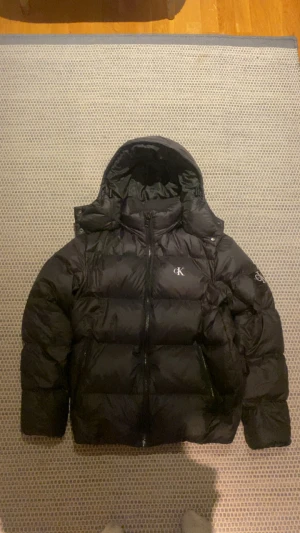 Svart pufferjacka från Calvin Klein - Svart pufferjacka från Calvin Klein med huva och dragkedja framtill. Jackan har CK-logga på bröstet och ärmarna, samt två sidofickor med dragkedja. Perfekt för kalla dagar med sin fluffiga och oversized look.