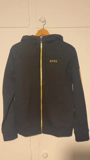 Svart hoodie från BOSS med gulddetaljer - Helt ny svart hoodie från BOSS med gulddetaljer på dragkedja, logga och längs ärmarna. Tröjan har huva med snörning, långa ärmar och två fickor framtill. Snygg och stilren design som sticker ut med sina kontrasterande detaljer.