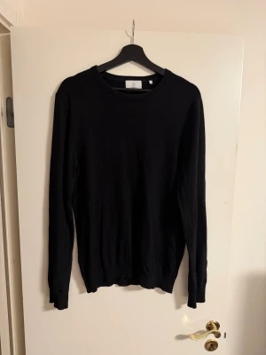 Svart tröja Bläck - En stilren svart stickad tröja från Bläck i storlek M. Tröjan har rund halsringning, långa ärmar och ribbade muddar vid ärmslut och nederkant. Perfekt för lager-på-lager och enkel att matcha med allt.