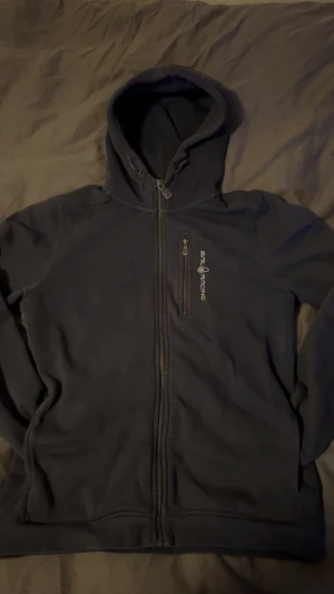 Mörkblå hoodie från Sail Racing - Snygg mörkblå hoodie från Sail Racing med hel dragkedja och huva. Tröjan har en bröstficka med dragkedja och logga tryckt på framsidan. Perfekt för dig som gillar sportig och clean stil.