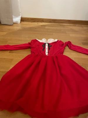 Röd klänning med vit krage från H&M - Supersöt röd klänning från H&M med vit krage och dekorativ svart rosett framtill. Klänningen har lång ärm, guldig knappdekor och en utställd kjol. Perfekt för dig som vill sticka ut med en klassisk men lekfull look.