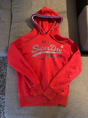 Röd Superdry hoodie med tryck - Riktigt snygg röd hoodie från Superdry med blått och vitt tryck på bröstet. Skick 7/10. Bara höra av dig vid funderingar😁