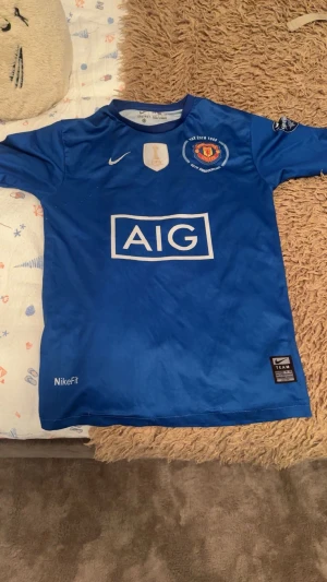 Blå Manchester United Ronaldo 7 tröja - Säljer en blå Manchester United fotbollströja från Nike med Ronaldo 7 på ryggen. Tröjan har AIG-logga på bröstet, klubbmärke och FIFA-märke framtill samt NikeFit Dri-FIT-material som andas. Kortärmad modell, perfekt för dig som älskar fotboll och United.