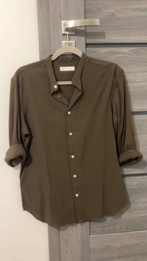 Brun henley-skjorta från ZARA - Brun henley-skjorta från ZARA med klassisk ståkrage och vita knappar framtill. Skjortan har långärmade ärmar som är snyggt uppvikta och en loose passform. Tillverkad i mjuk bomull som känns skön mot huden. Perfekt för dig som gillar enkel och stilren design.