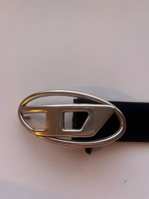 Svart skärp från Diesel med silverfärgat spänne - Snyggt svart bälte från Diesel i 100% skinn med ett stort, ovalt silverfärgat spänne med Diesel-logga. Perfekt accessoar för att lyfta din outfit med en cool detalj. Bältet är stilrent och passar till jeans eller byxor för en trendig look. Köpt från nk så garanterat äkta!