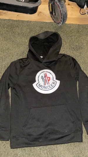 Svart Moncler hoodie med stor logga - Svart hoodie från Moncler med stor, vit och röd logga på bröstet. Tröjan har huva, lång ärm och en klassisk magficka framtill. Perfekt för dig som gillar streetwear och vill ha en snygg, enkel look med tydlig branding.