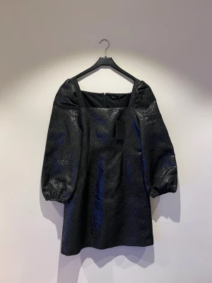 Glittrig svart kortklänning Stockh LM - Supersnygg svart klänning från Stockh LM med glittrigt tyg och puffiga långärmar. Klänningen har rak passform, fyrkantig urringning och ballongärmar med resår vid ärmslut. Perfekt för dig som vill sticka ut med en trendig look. Perfekt till nyår. 