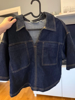 Mörkblå denimskjorta  - Snygg mörkblå denimskjorta från Kappahl i storlek XS. Skjortan har korta ärmar, två stora bröstfickor och kontrastsömmar som ger en cool look. Modellen är loose fit med v-ringad hals och klassisk krage. Perfekt att styla med jeans eller kjol.