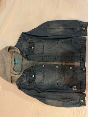 Jeansjacka med grå luva SWEET SKTBS - Snygg jeansjacka från SWEET SKTBS i blå denim med fastsydd grå huva. Jackan har knappar framtill, två bröstfickor med lock och sidofickor. Märkespatch nertill och coola detaljer på knapparna. Mindre hål (6 mm~ ) i ena fickan men inget som märks för övrigt, se bild ovan. 